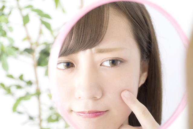 手鏡の中の自分を見つめ、指先で肌の赤みや痛みの状態を確認している女性の様子｜アーバンコスメ