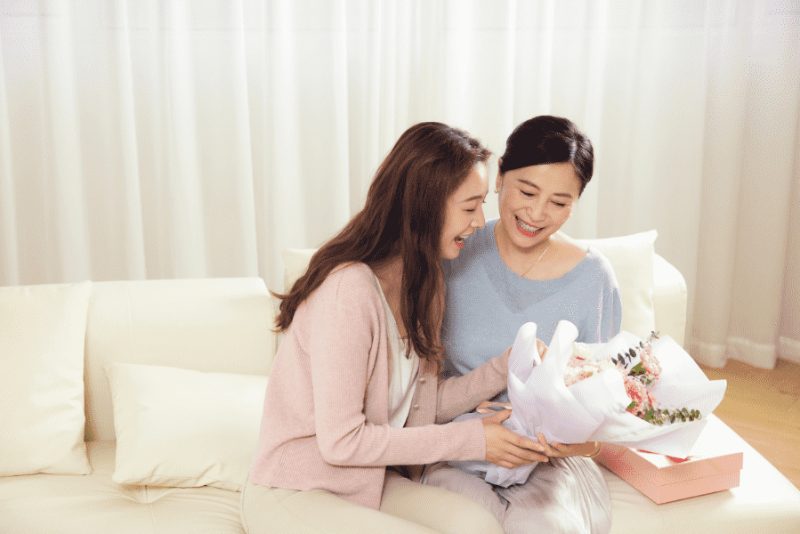 母の日のプレゼントとして贈られた花束を一緒に眺め、温かい光が差し込むリビングで仲睦まじく笑顔を交わす母娘の幸せなひとときの様子|アーバンコスメ