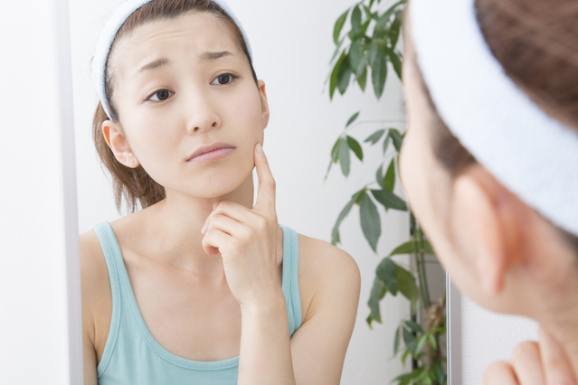 鏡の前で肌に指を添え、テカリや化粧崩れが気になるのにお肌を触ると硬く感じたり、毛穴が目立ったり、なかなか改善しない春先の不安定な肌コンディションを深刻そうに見つめている女性の様子｜アーバンコスメ