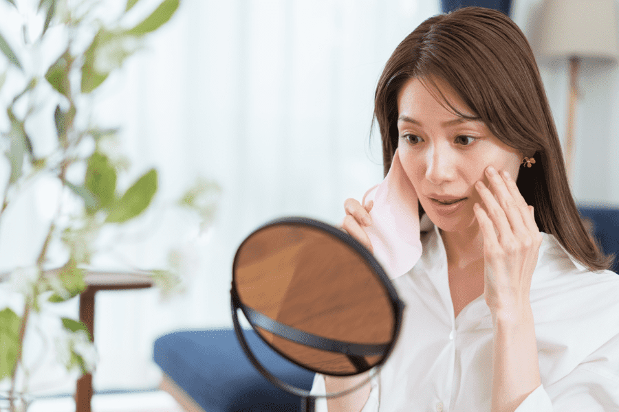 鏡を見ながら自分の肌の状態を手で触って確認し、表面のテカリとは裏腹に肌の奥に感じるつっぱりや違和感に、インナードライの兆候を感じている女性の様子｜アーバンコスメ