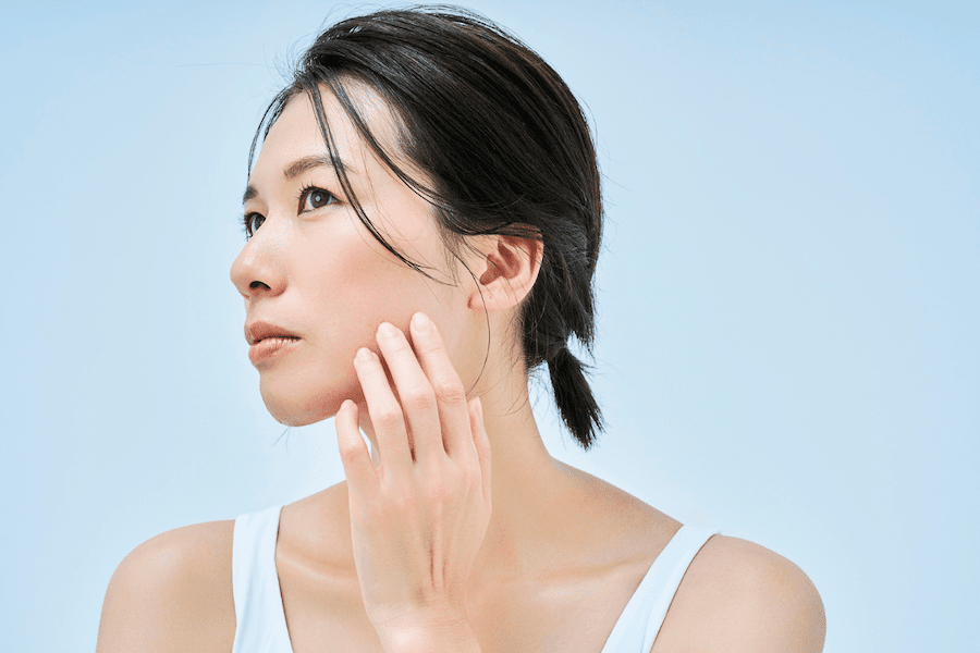 初夏の明るい光の下で、気温の上昇とともに気になり始めたたるみ毛穴やハリ不足を鏡越しにチェックし、内側から引き締まった上向き肌へのケアを意識している女性の様子｜アーバンコスメ