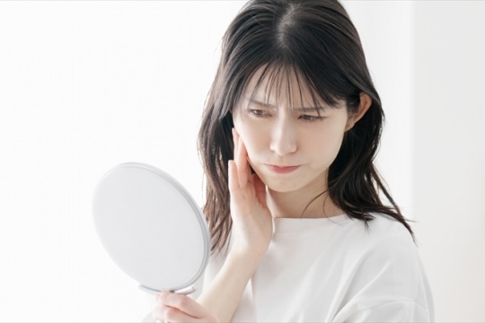 鏡の前で指先を頬にそっと添え、季節の変わり目や乾燥によって突然現れた大人の吹き出物や硬くなった角質による肌の違和感を静かに確認している女性の様子｜アーバンコスメ